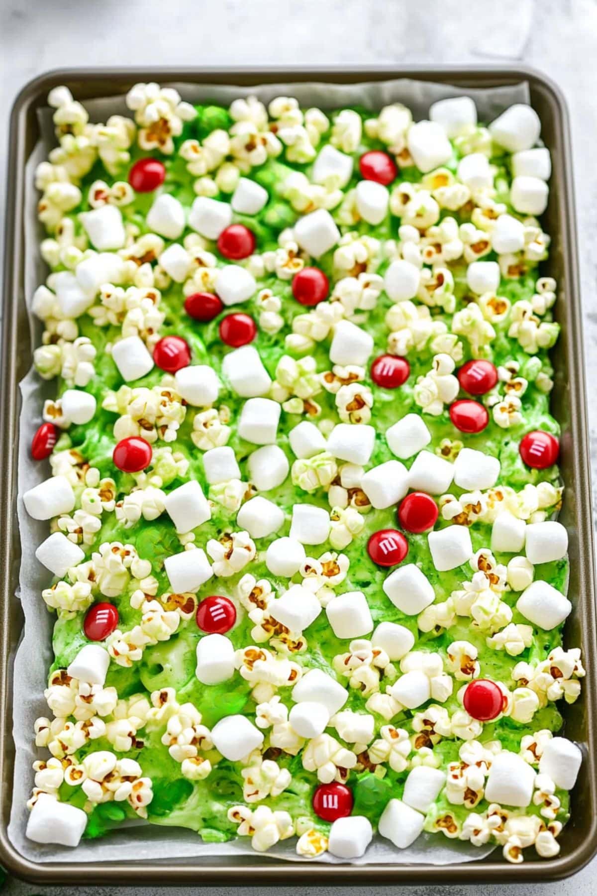 Green colored popcorn spread in a baking sheet with parchment paper with red candies and mini marshmallow on top.