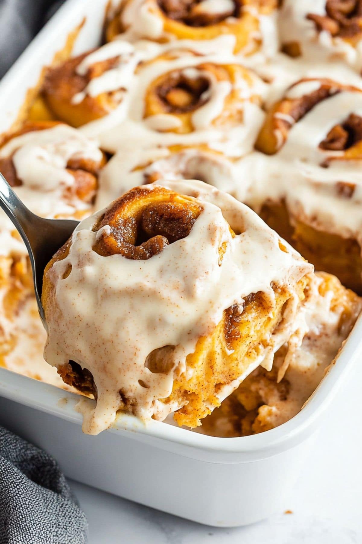 Pumpkin cinnamon roll with cream cheese frosting lifted by a pie ladle from a white baking dish.