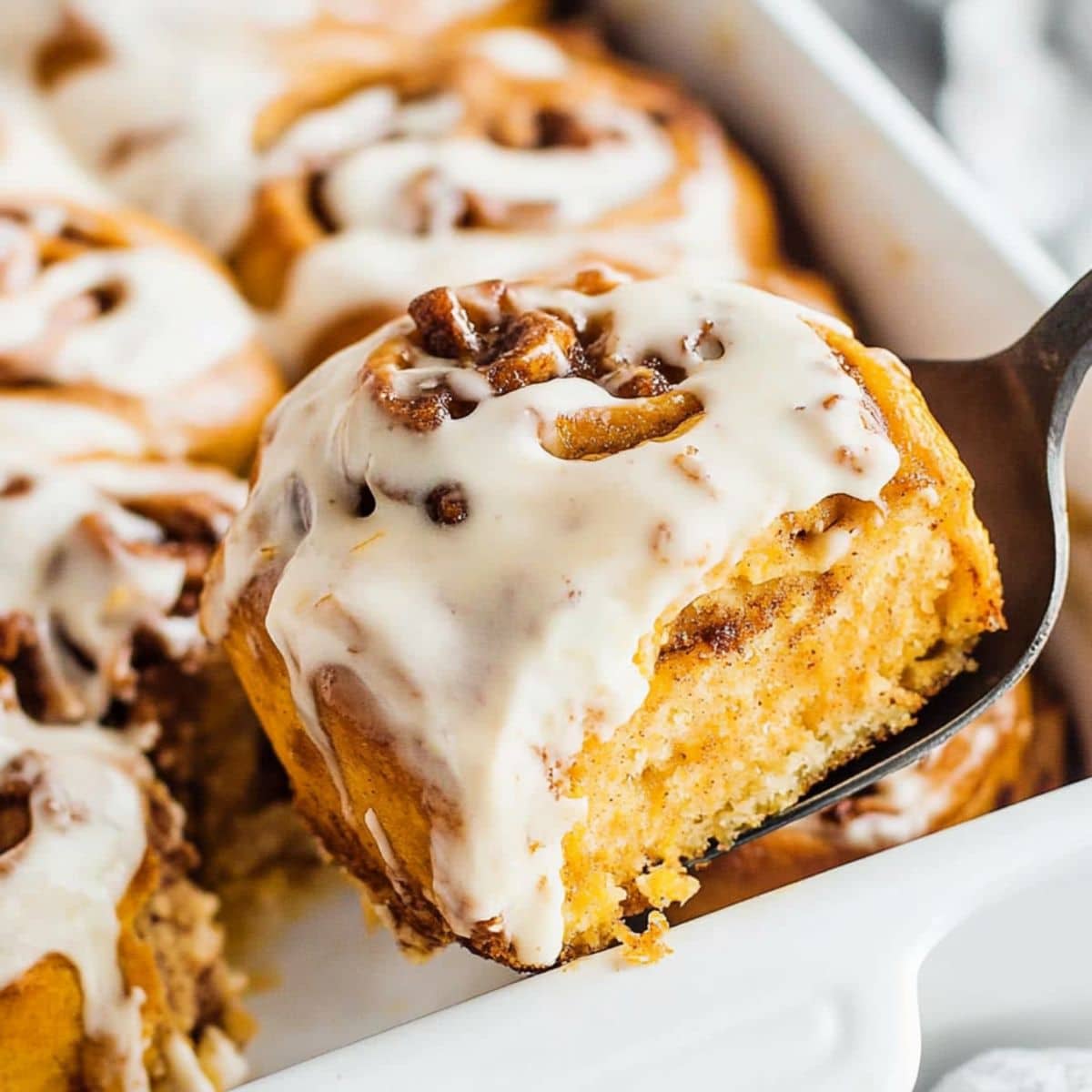 Pie ladle lifting a serving slice of pumpkin cinnamon rolls from baking dish.