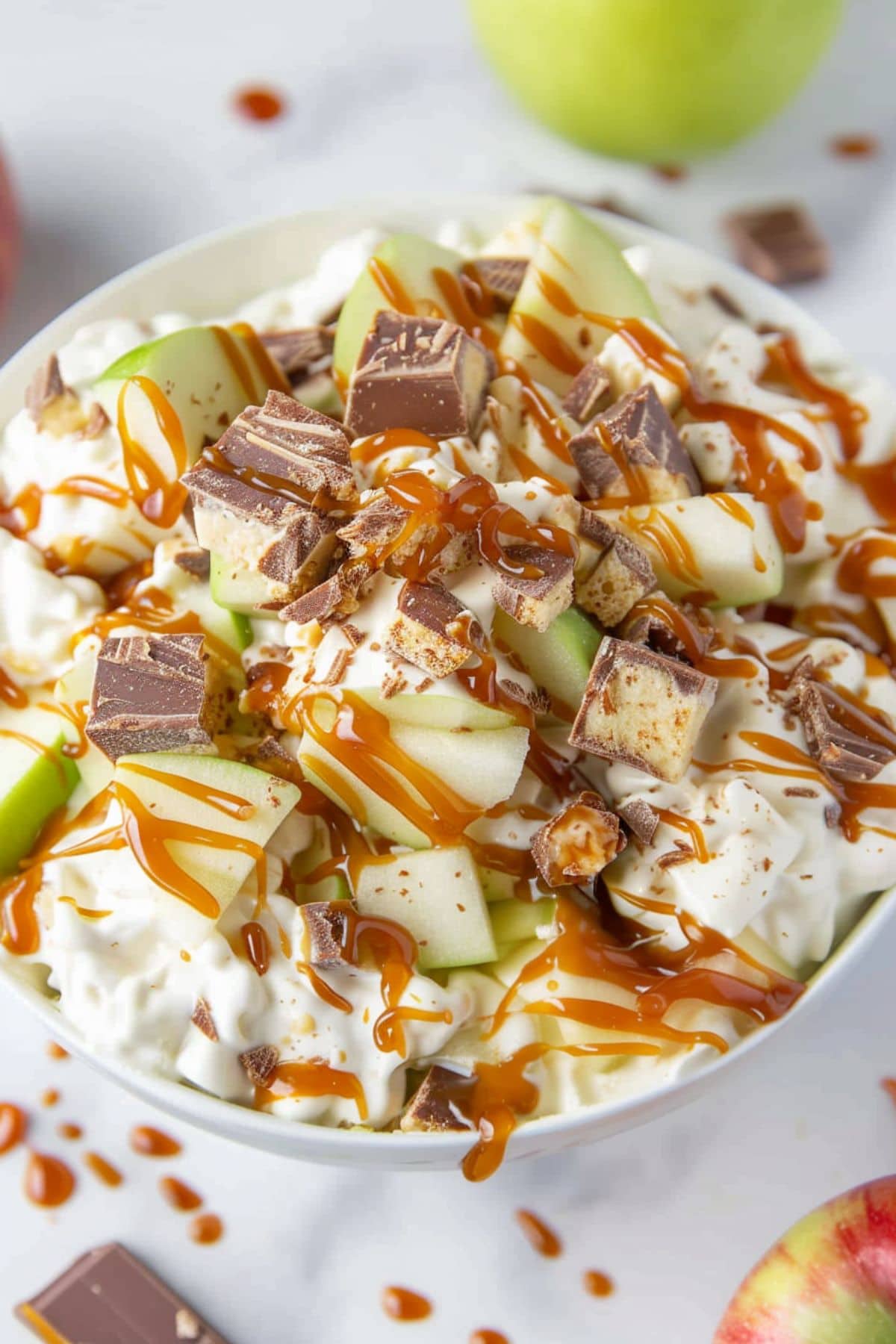 Chopped Snickers candy bars and green apples in creamy pudding drizzled with caramel sauce drizzled on top, served in a white bowl.