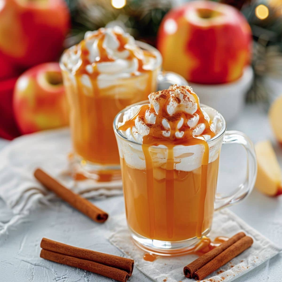 Starbucks caramel apple cider drinks topped with whipped cream drizzled with caramel syrup served in glass mugs.