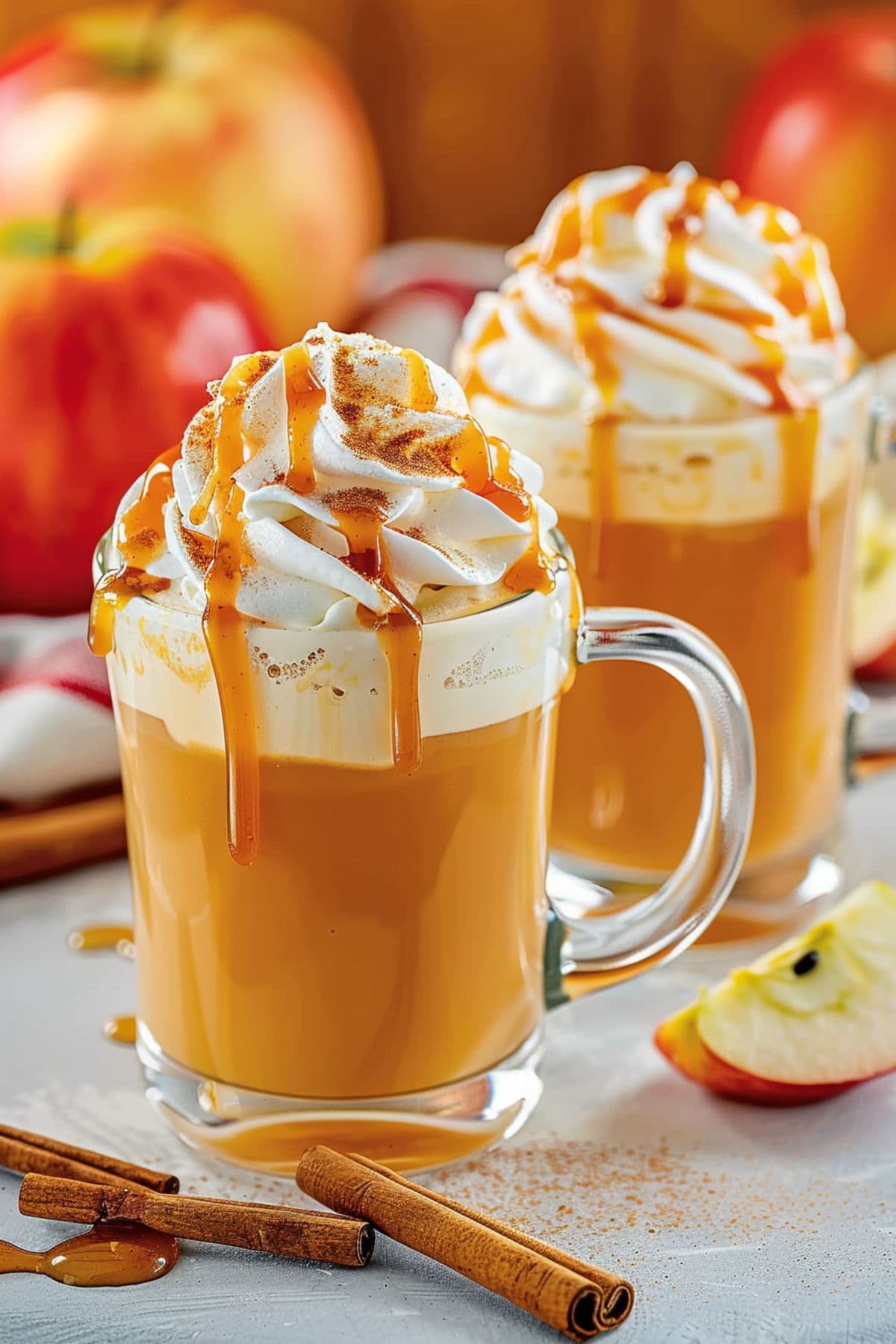 Two glass mugs with apple cider drink with whipped cream drizzled with caramel sauce.