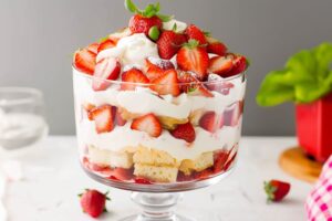 Strawberry shortcake assembled inside a large glass trifle dish garnished with fresh strawberries on top.