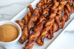 Homemade crunchy bacon twists in a platter, served with a bowl of brown sugar.