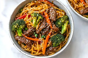 A close-up of beef and broccoli lo mein with crispy carrots and garnished with sesame seeds in a bowl.