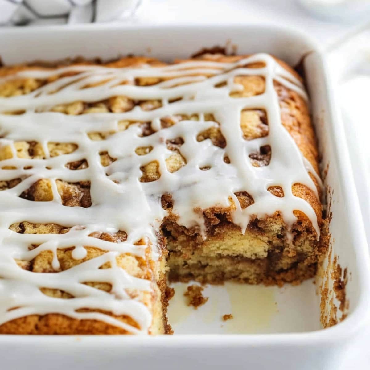 Square slice portion missing on a cinnamon roll cake baked in a baking dish drizzled with white glaze on top.