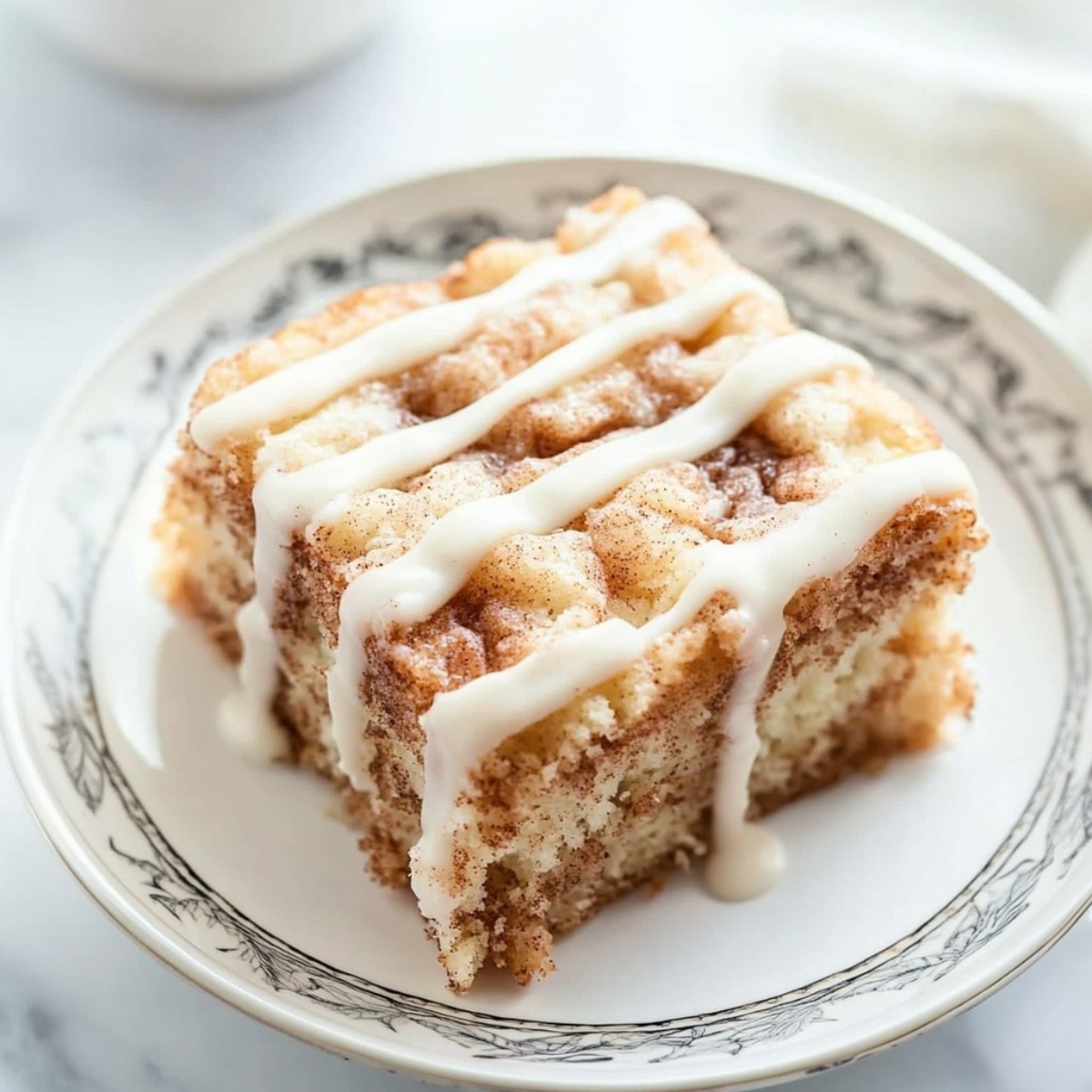 Square slice of cinnamon roll coffee cake served in a saucer drizzled with sugar glaze on top.