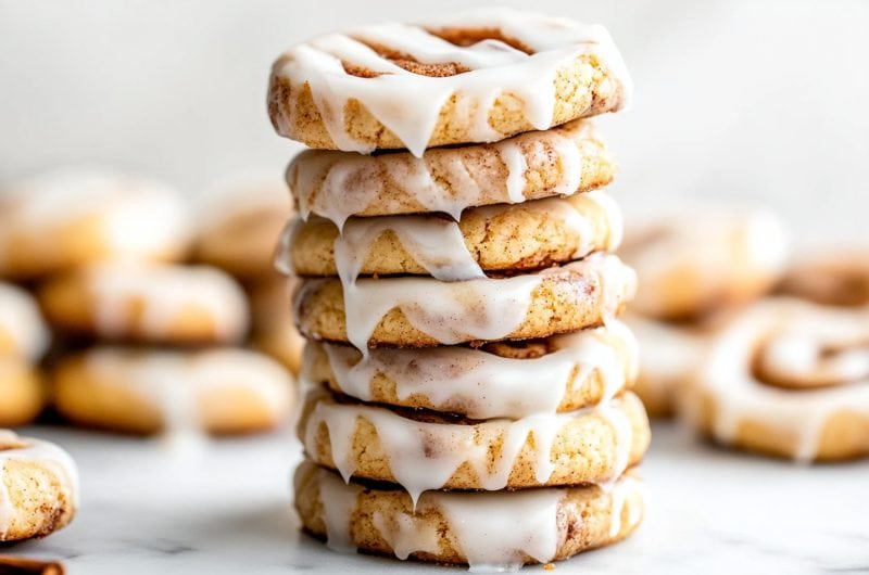 A stack of cinnamon roll cookies drizzled with glaze sitting on a marble table.