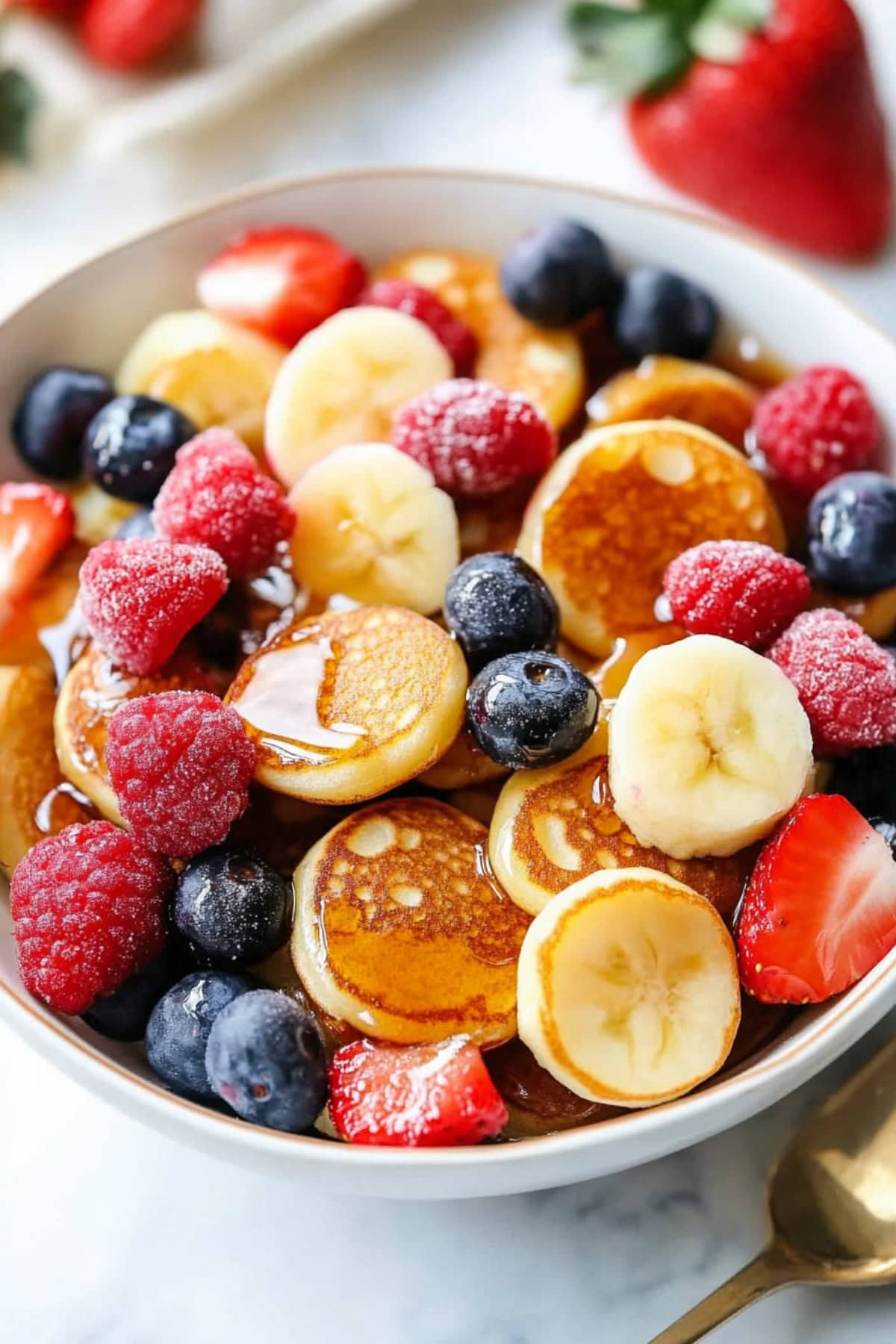 Mini pancake cereal with berries and syrup in a white bowl.