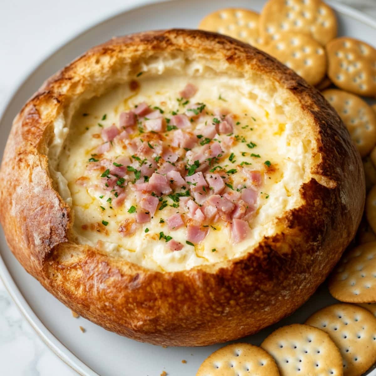 Loaf bread hollowed in the middle filed with cheese dip with ham and green onions served with a platter of crackers.