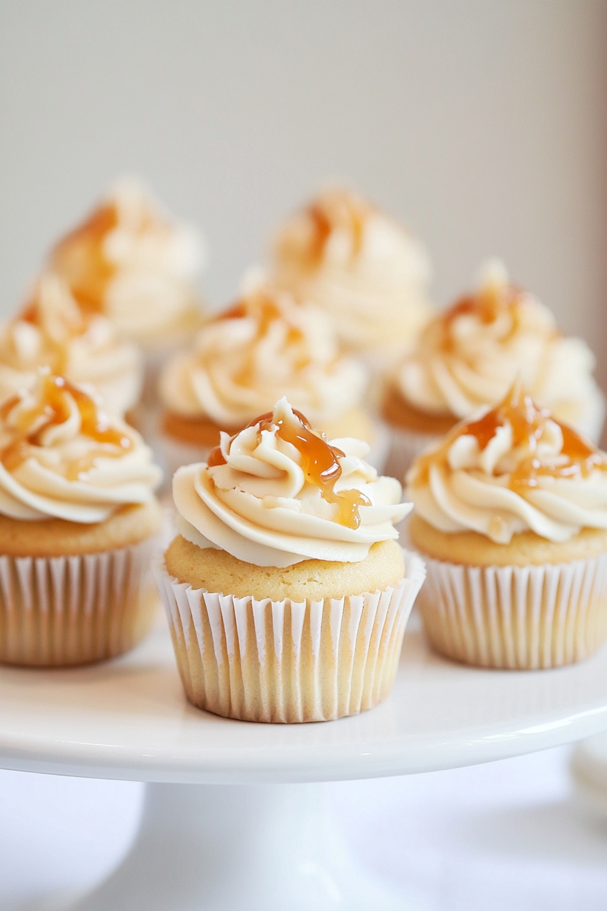 Vanilla cupcakes topped with vanilla frosting and drizzled with caramel on a white cake stand