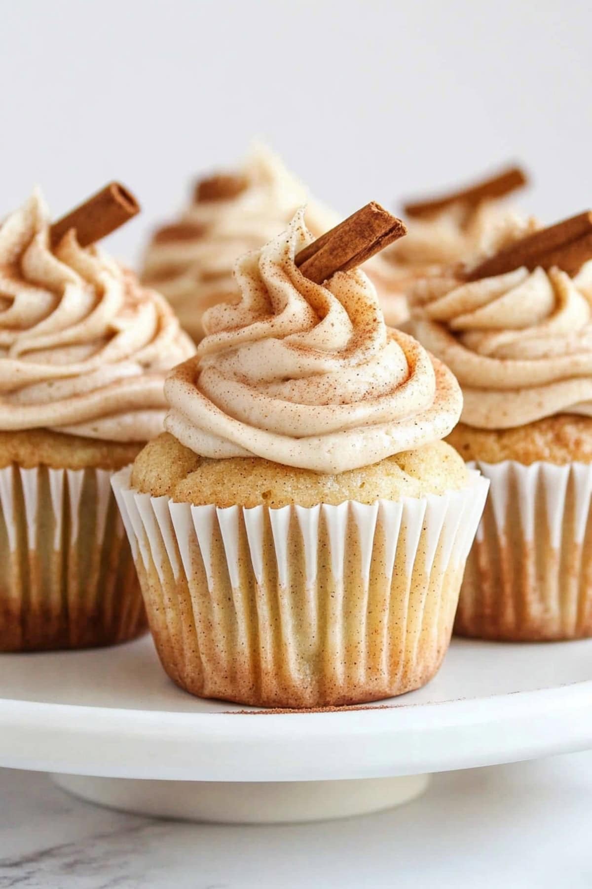 Snickerdoodle cupcakes with creamy frosting garnished with cinnamon sticks, sitting on a cake stand.