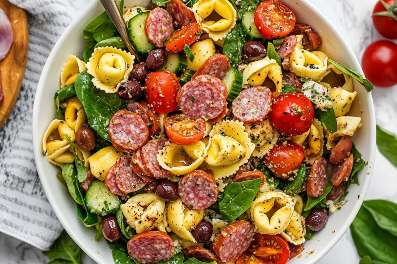 Festive tortellini pasta with tomatoes, cucumber, red onion, olives, mozzarella, salami, Parmesan, spinach, and basil tossed in dressing served in a white bowl.