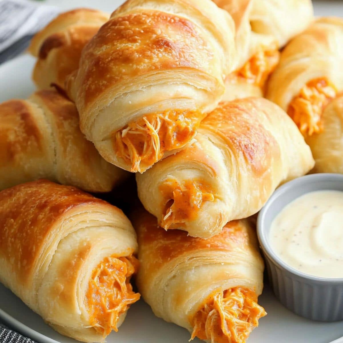 Bunch of crescent rolls with shredded buffalo chicken filling inside served in a white plate with ranch dressing on the side.