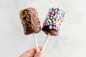 Hand holding two sticks with chocolate covered marshmallows covered with sprinkles.