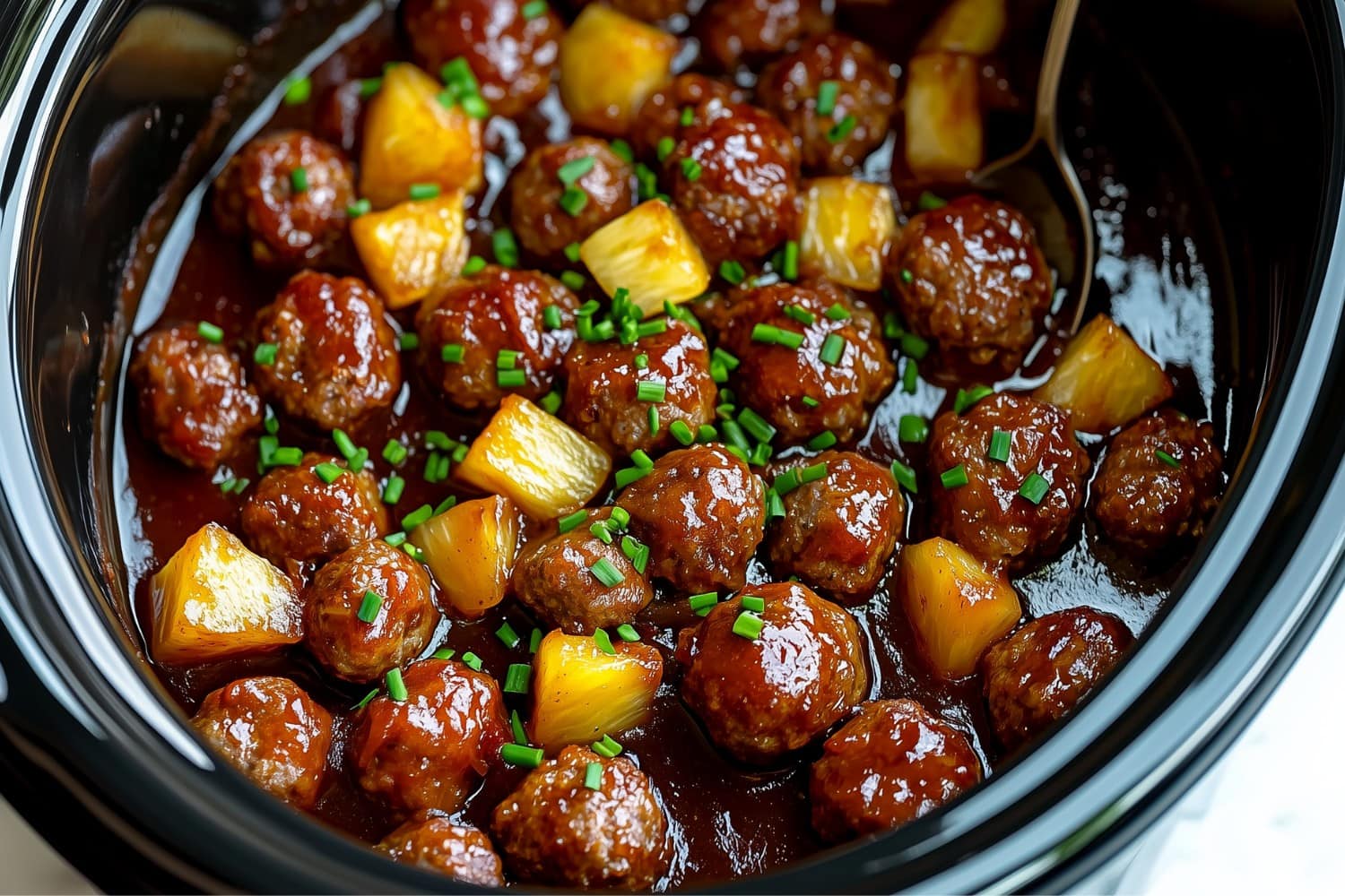 A close-up of Crockpot Hawaiian meatablls in rich barbecue sauce, with pineapple chunks and chopped chives.