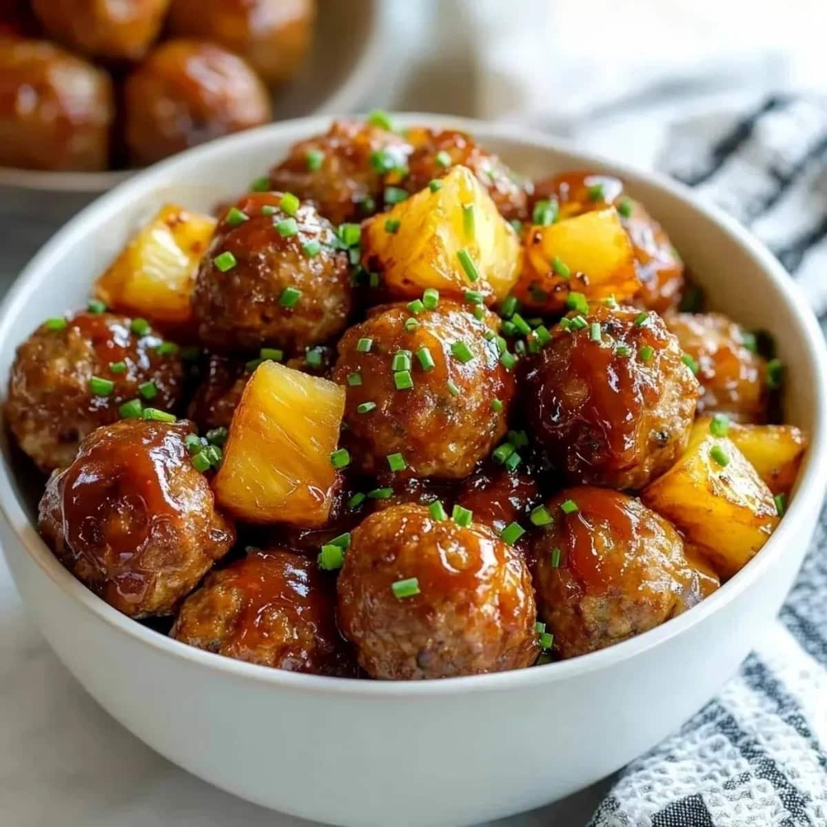 A bowl of Hawaiian meatballs with barbecue sauce and pineapple chunks, garnished with chopped chives.