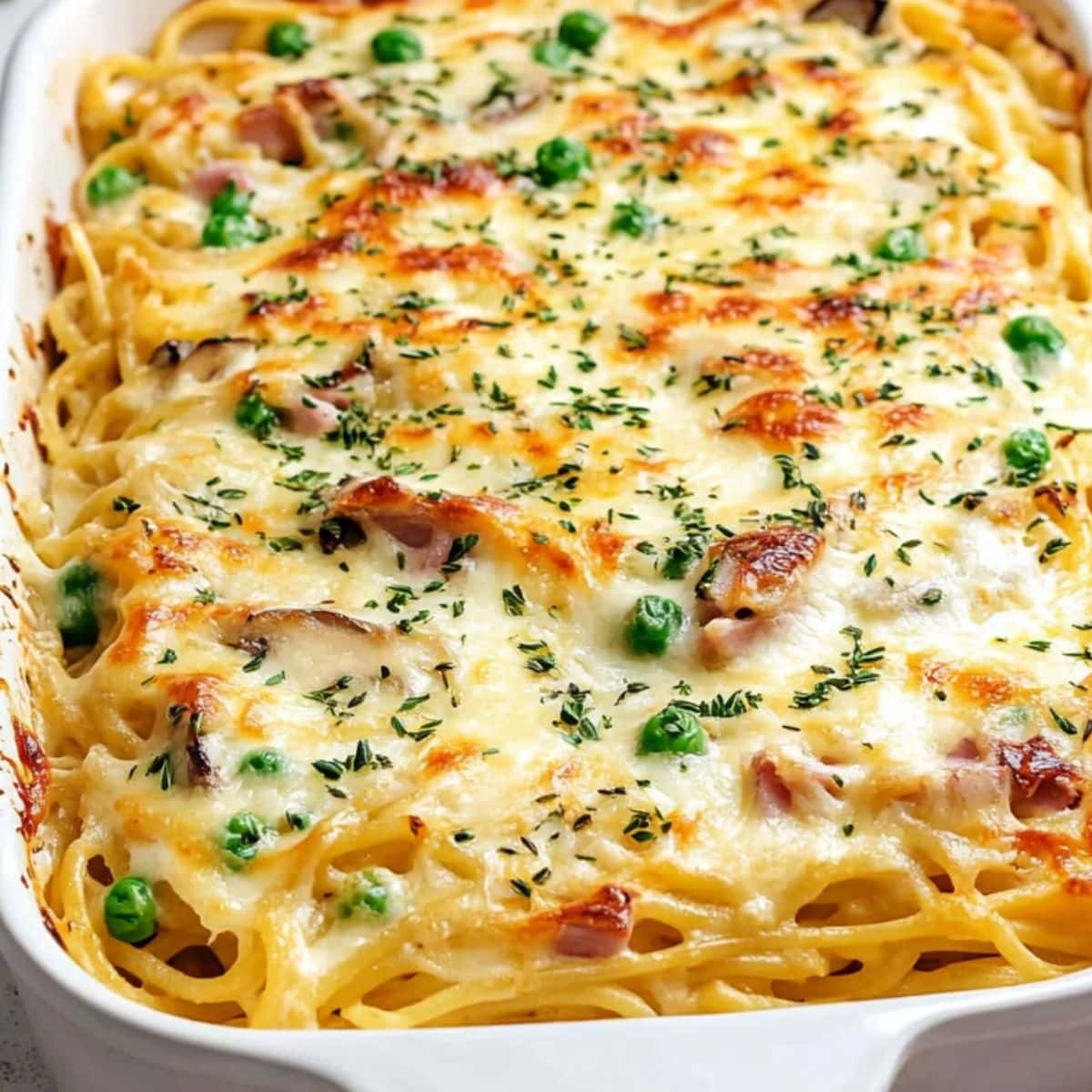 Baking dish with Baked spaghetti, ham, mushrooms, and peas with creamy and cheesy sauce topped with melted cheese.
