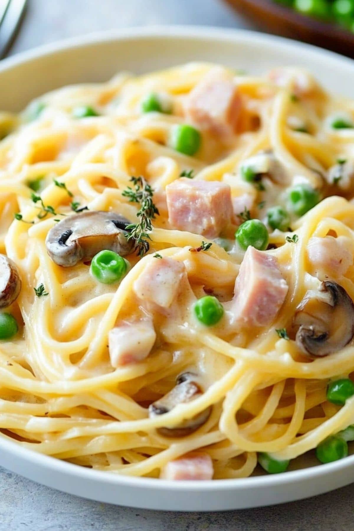 A serving of Spaghetti, ham, mushrooms, and peas with creamy and cheesy sauce from white plate.