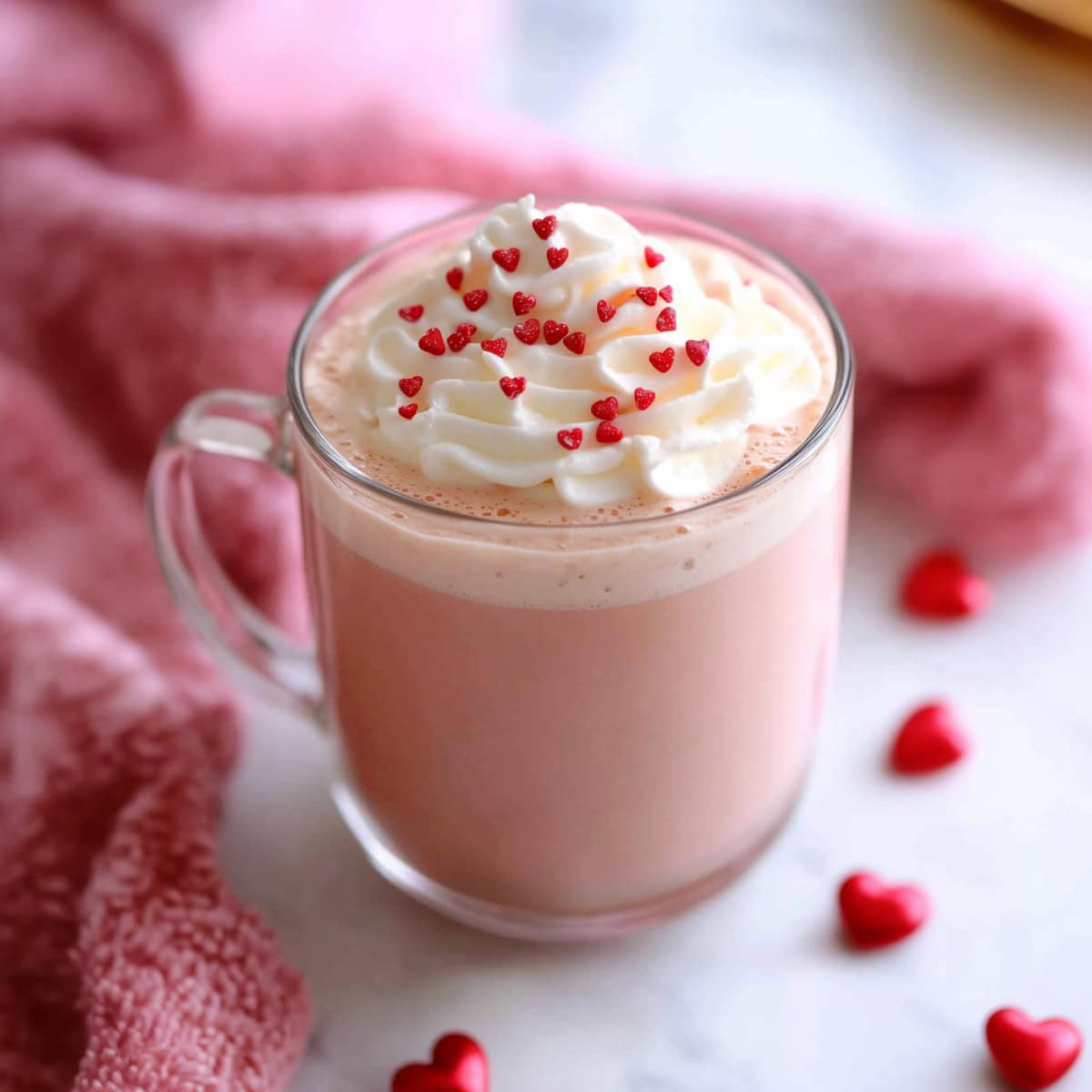 A glass mug of pink hot chocolate topped with whipped cream and heart-shaped candies.