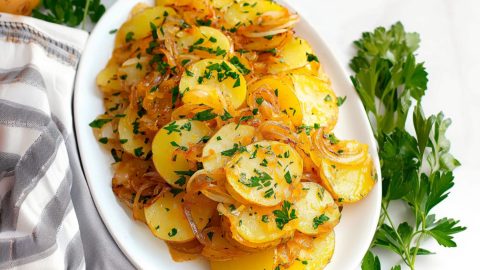Sliced potatoes with buttery sauce and caramelized onions served in a white platter with chopped parsley.