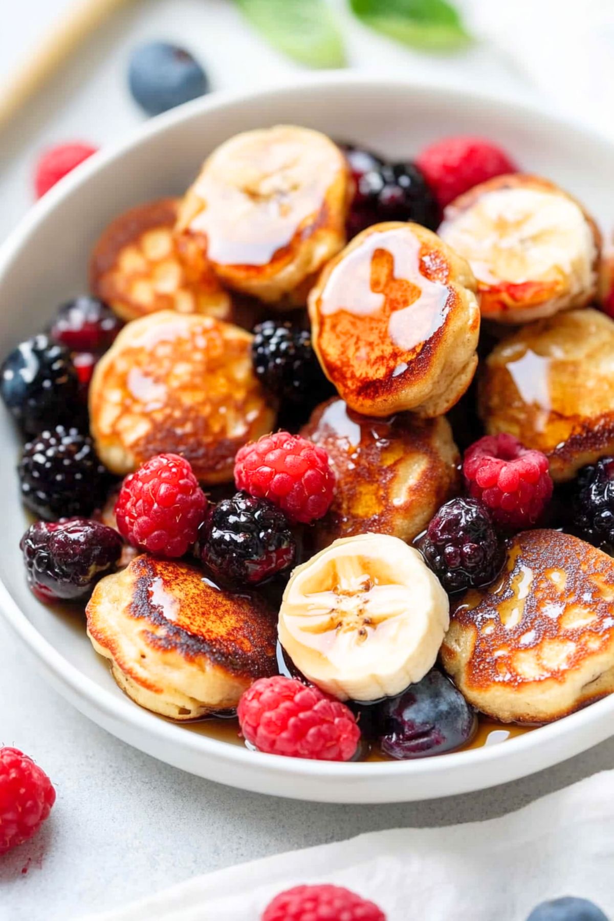 Bunch of banana pancake bites served with berries drizzled with maple syrup in a bowl.