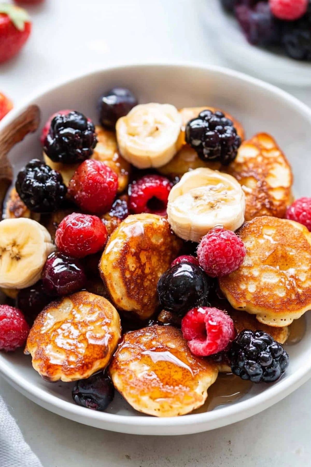 Mini banana pancake bites served in a white plate with berries and drizzled with maple syrup.