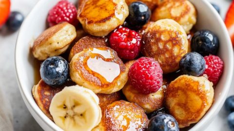 Mini banana pancake bites served in a white bowl with fresh berries and drizzle of maple syrup.