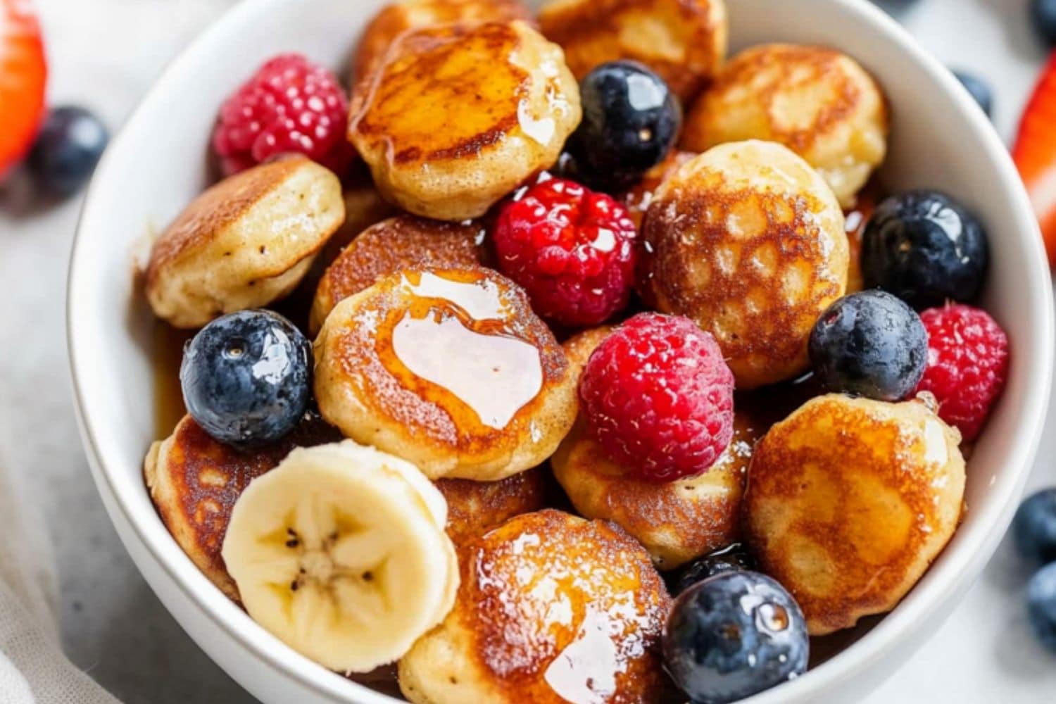 Mini banana pancake bites served in a white bowl with fresh berries and drizzle of maple syrup.