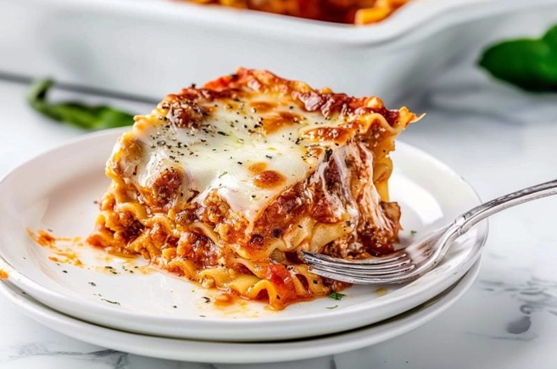 Lasagna slice serving in a white plate with fork on the side.