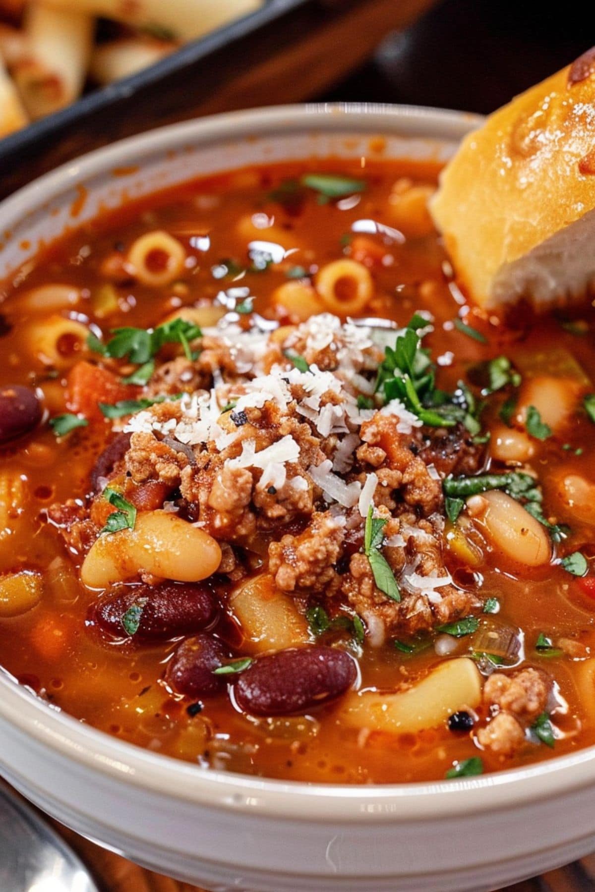 Pasta e Fagioli with beans beans, pasta, and tomato broth served in a white bowl with a piece of bread on the side garnished with sprinkle of parmesan cheese.