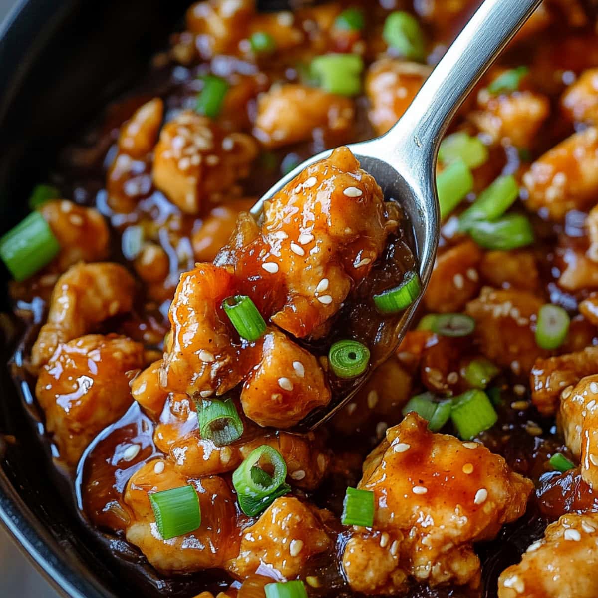 A spoonful of saucy sesame chicken with chopped green onions in a saucepan.