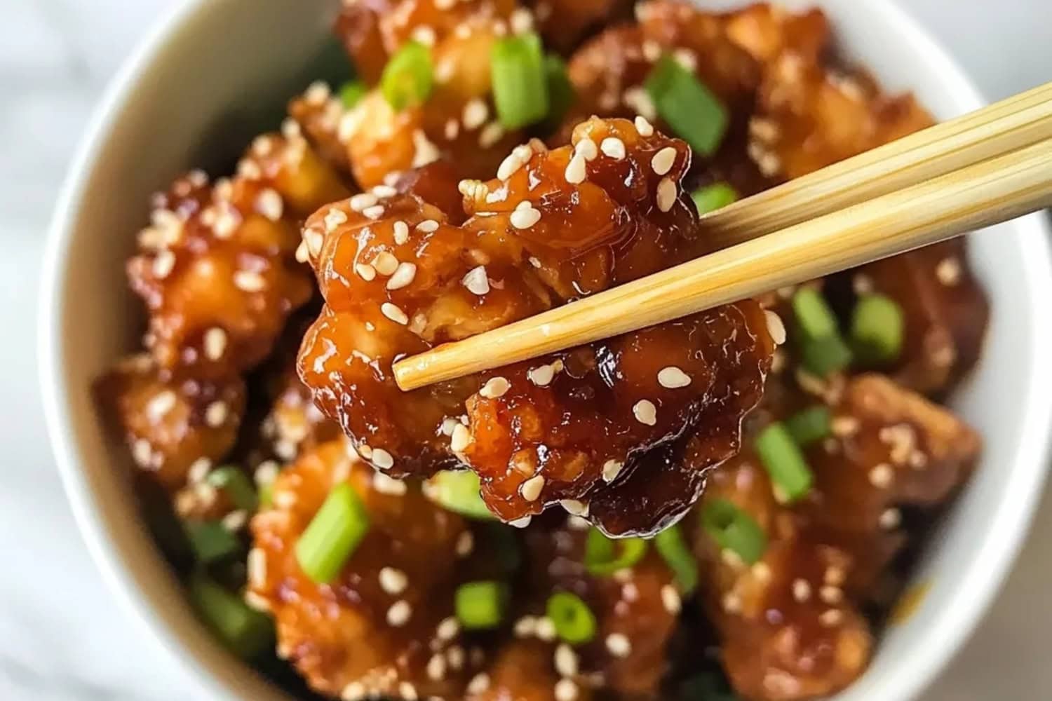 Close-up of sesame chicken with green onions in chopsticks.