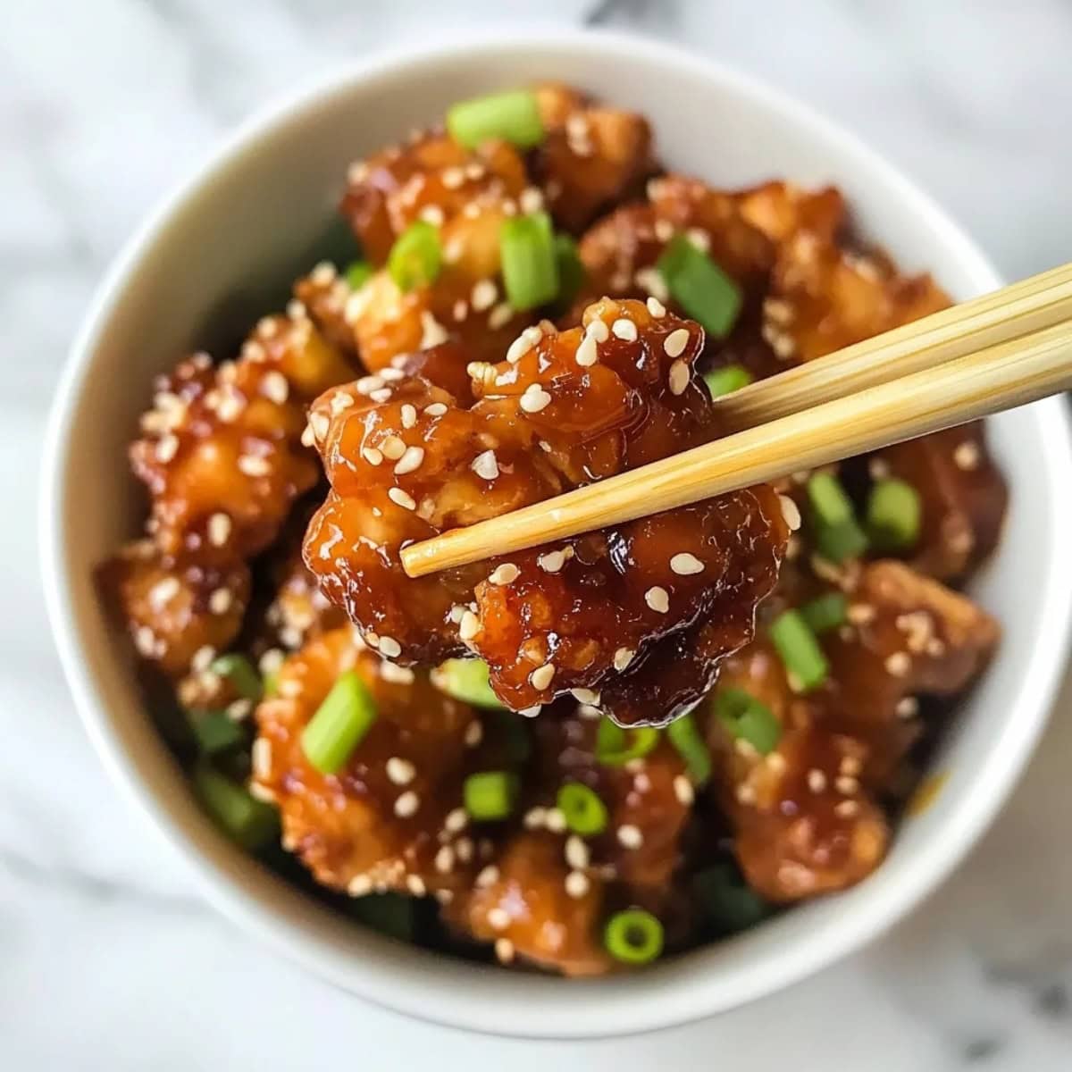 Chopsticks holding a piece of sesame chicken with chopped green onions.