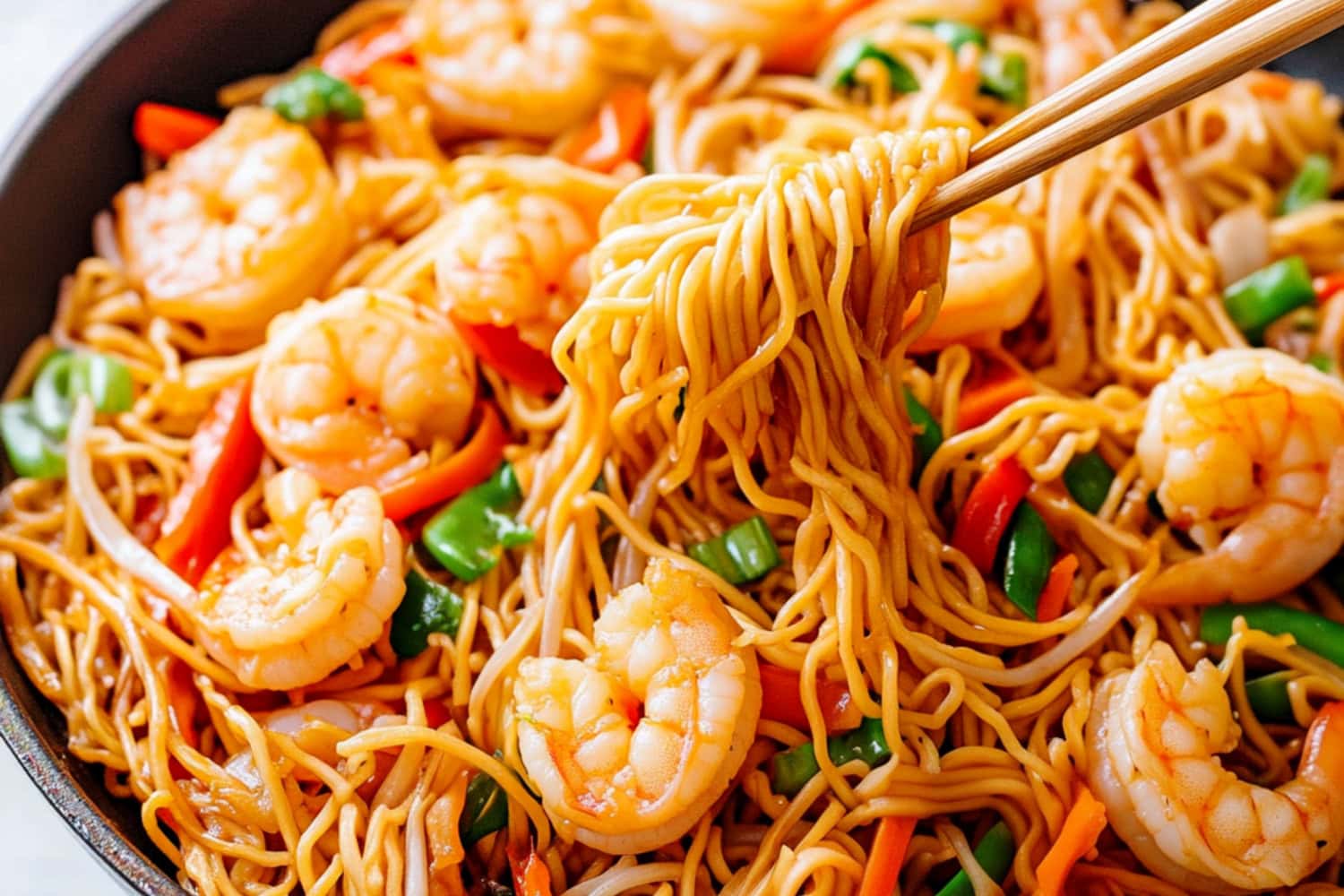 Chopsticks lifting shrimp chow mein noodles from a skillet, with shrimp, bell peppers, carrots and green onions.