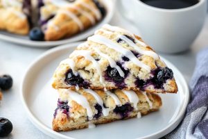 Two servings of blueberry scones with drizzle of sugar glaze served in white plate with cup of coffee on the side.