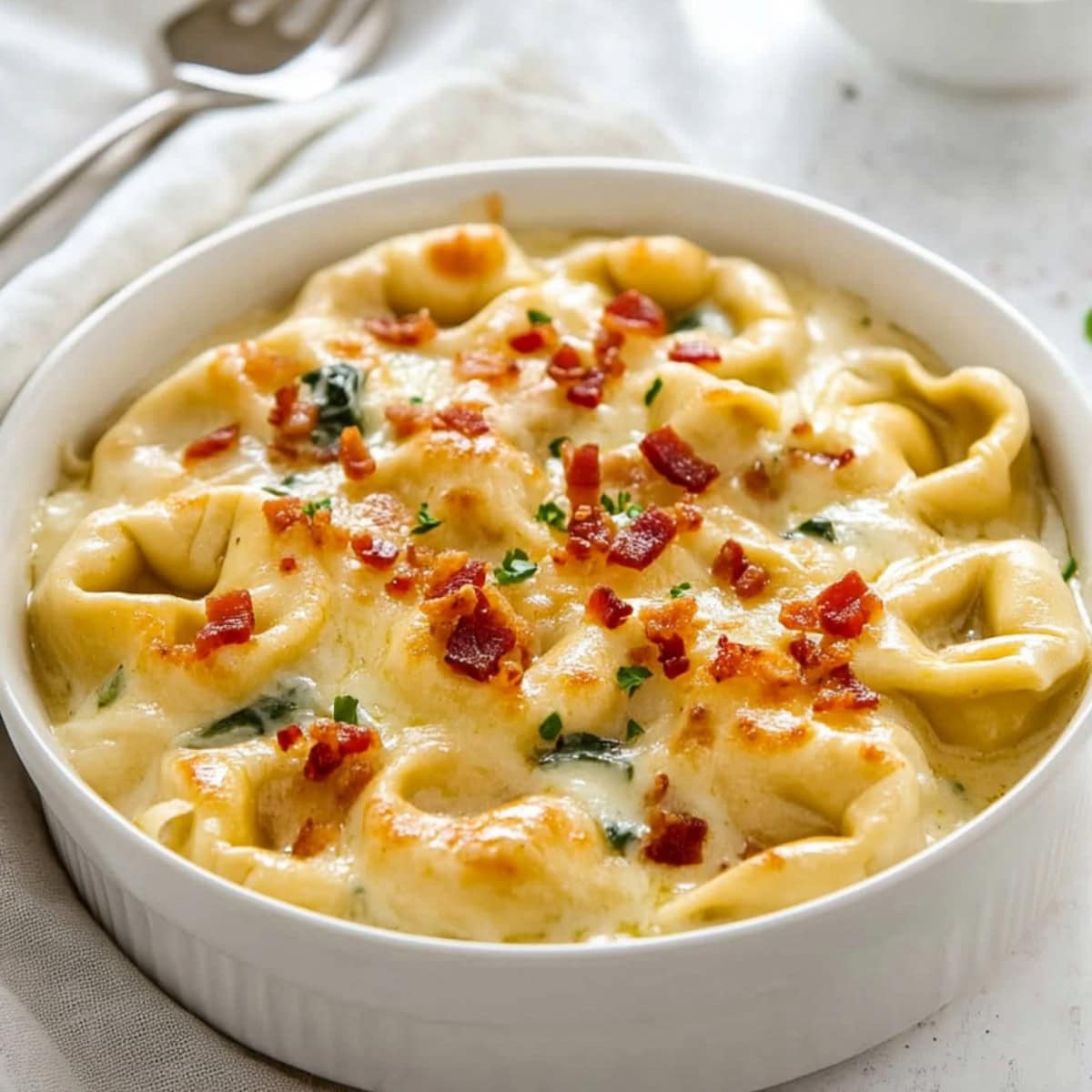 White bowl with a serving of cheesy tortellini al forno with creamy sauce, chopped bacon and spinach.