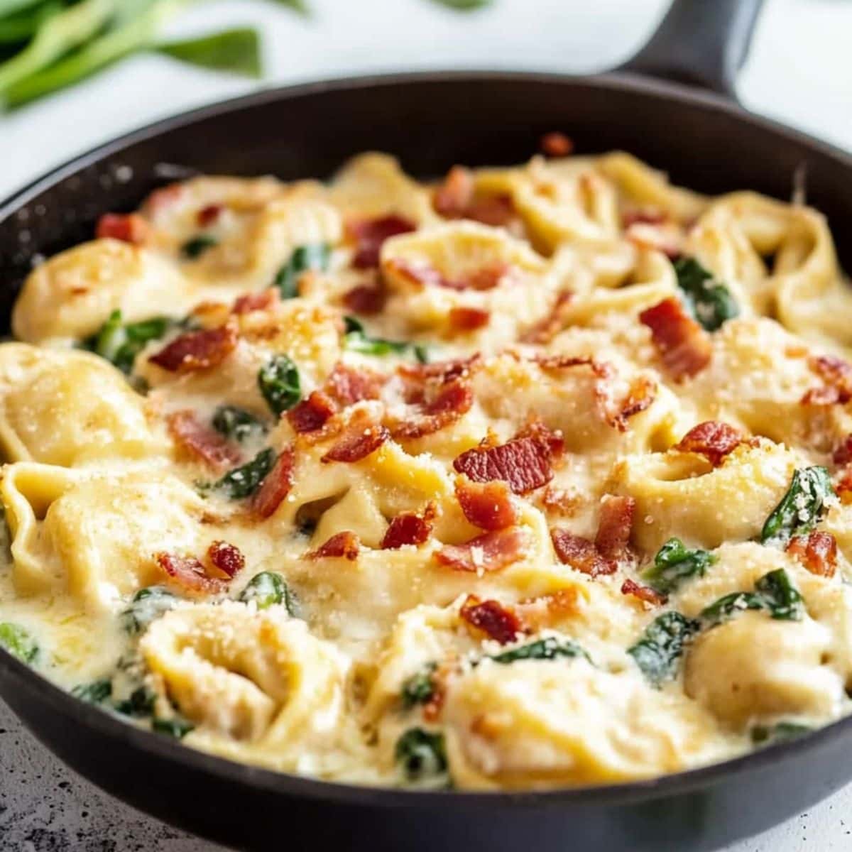 Skillet pan with tortellini pasta cooked in creamy and cheesy sauce with bacon and spinach.