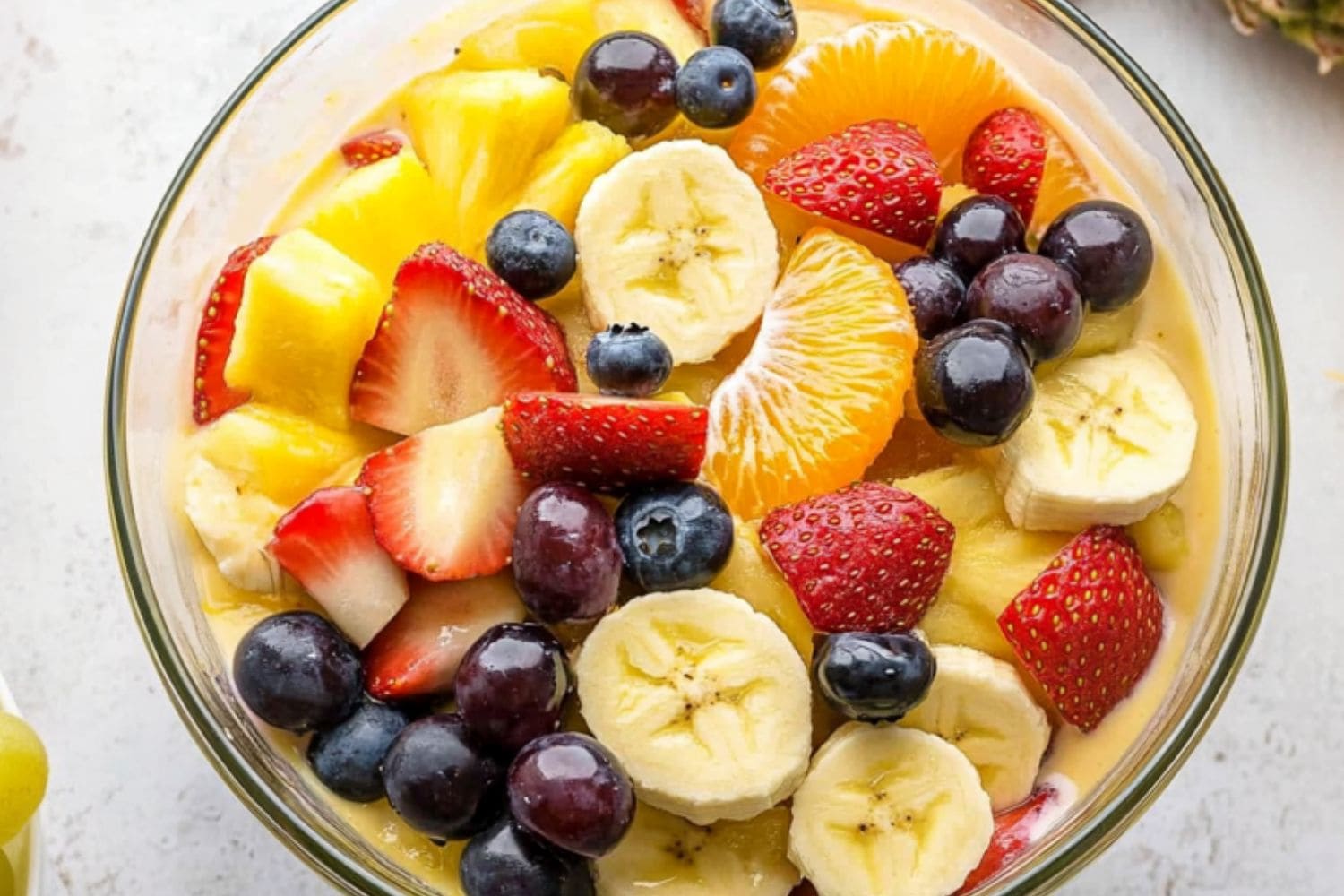 Vanilla pudding fruit salad with pineapple chunks, mandarin oranges, bananas, strawberries, blueberries, and grapes tossed in a glass bowl.