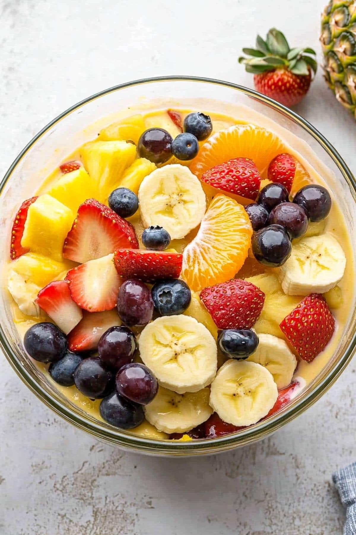 Large glass bowl with mix of pineapple chunks, mandarin oranges, bananas, strawberries, blueberries, and grapes and vanilla pudding mix.