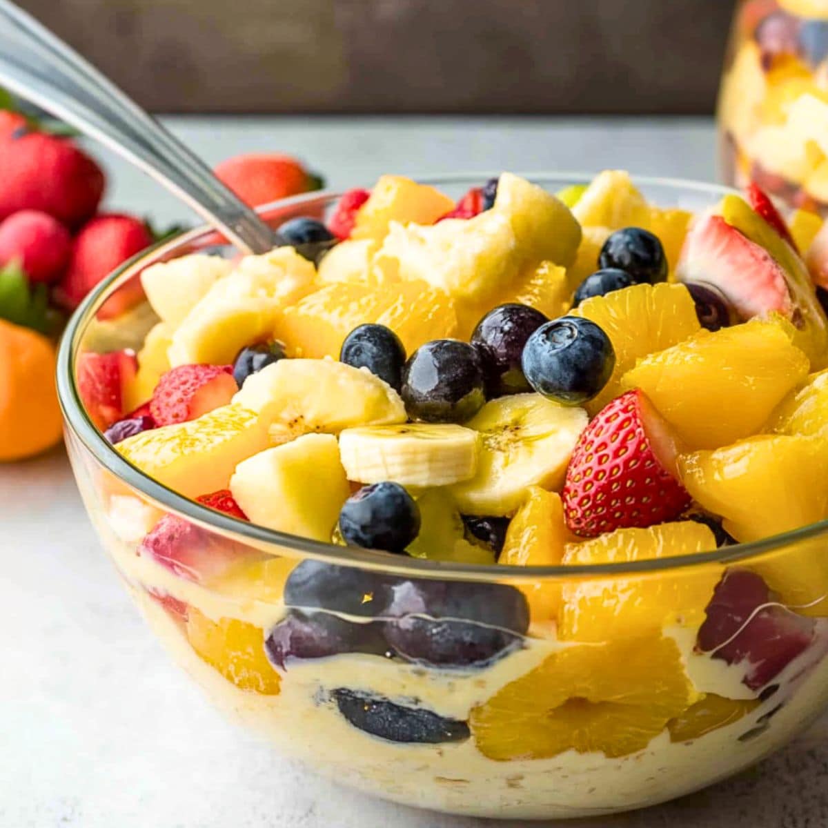 Large glass bowl wit fruit salad made with pineapple chunks, mandarin oranges, bananas, strawberries, blueberries, and grapes tossed in vanilla pudding.