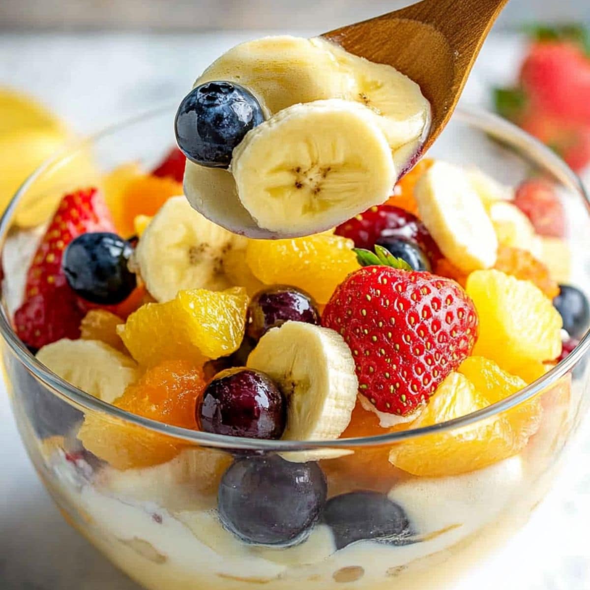 Wooden spoon tossing vanilla padding fruit salad in a glass mixing bowl.