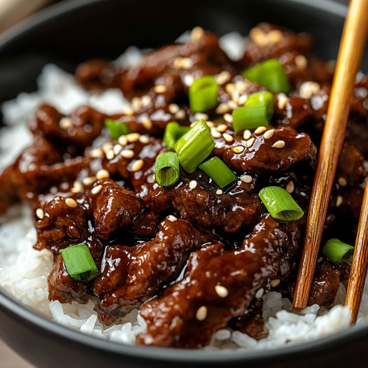 Beef teriyaki served on top of white rice in a black bowl with a sprinkle of sesame seeds and chopped onions.