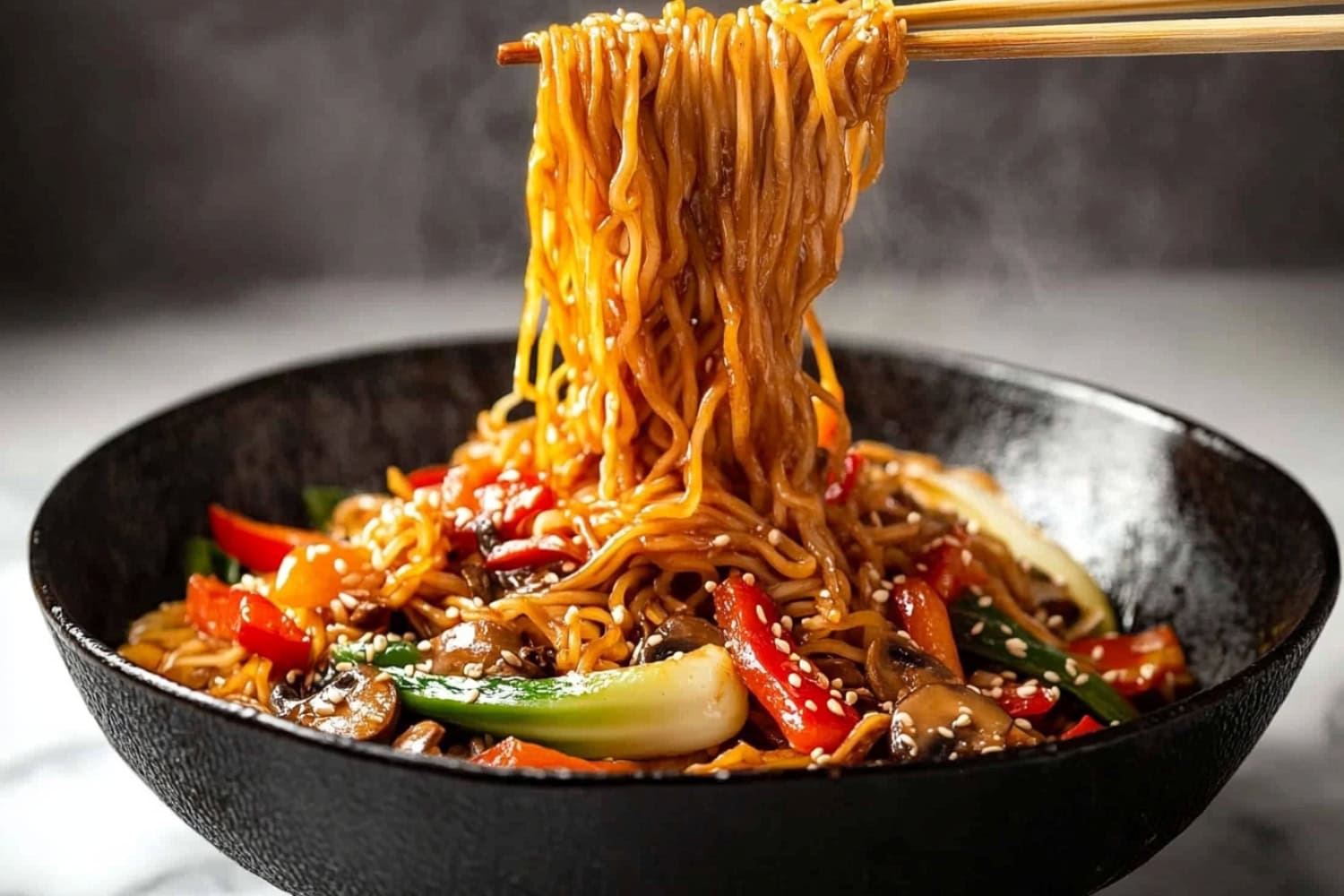 A close-up of chopsticks lifting some lo mein noodles from a bowl, surrounded by mixed vegetables and savory sauce.