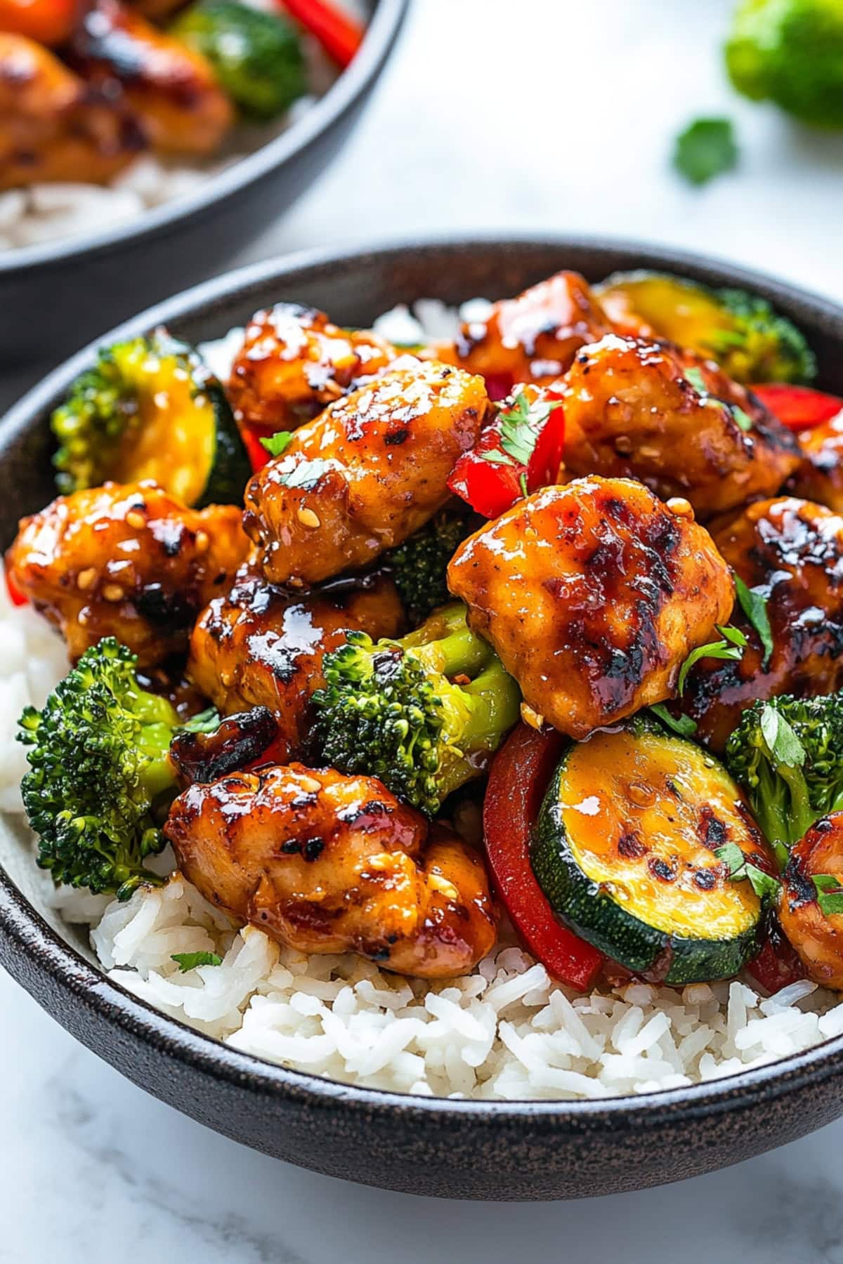 Bite sized chicken pieces cooked in Hunan sauce with broccoli florets, chopped red bell pepper, and zucchini served on top of white rice in a white bowl.