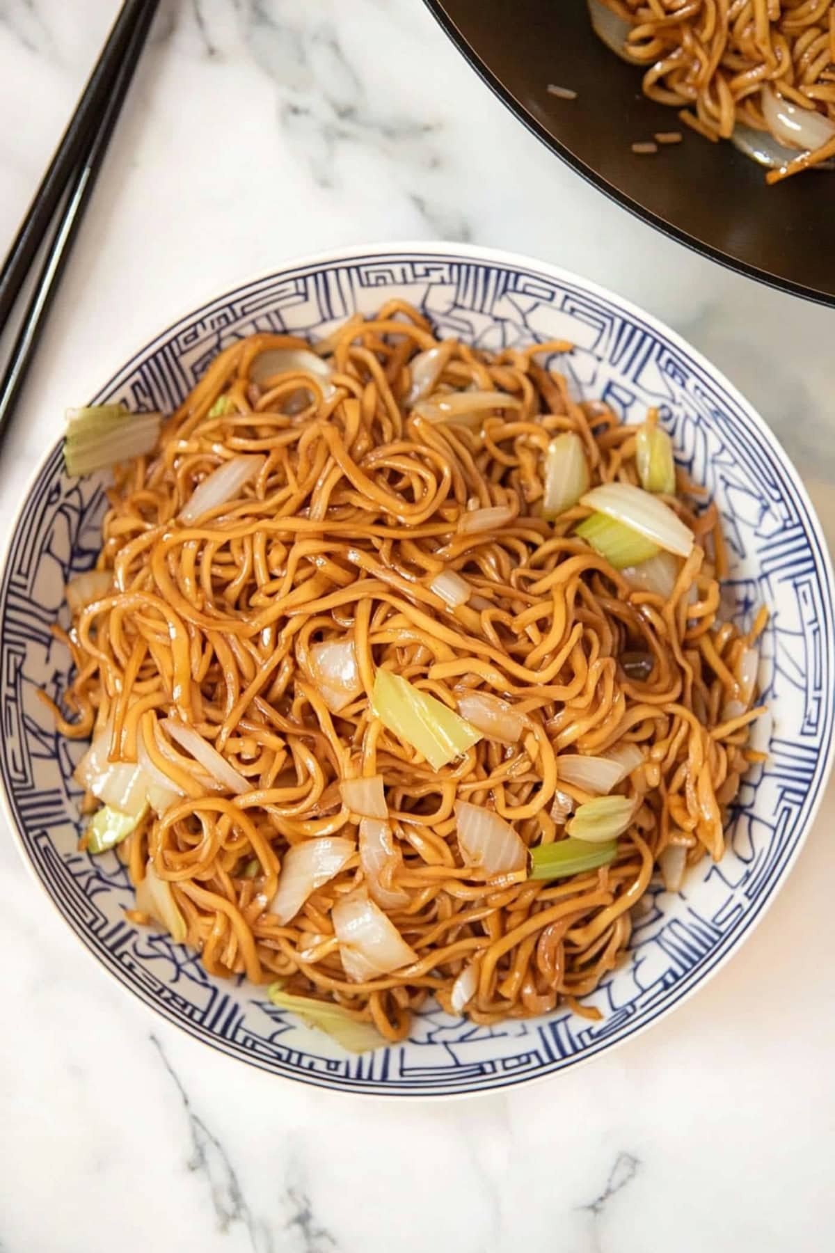 Chow mein noodles served in a Chinese bowl next to a skillet pan with the rest of the dish.
