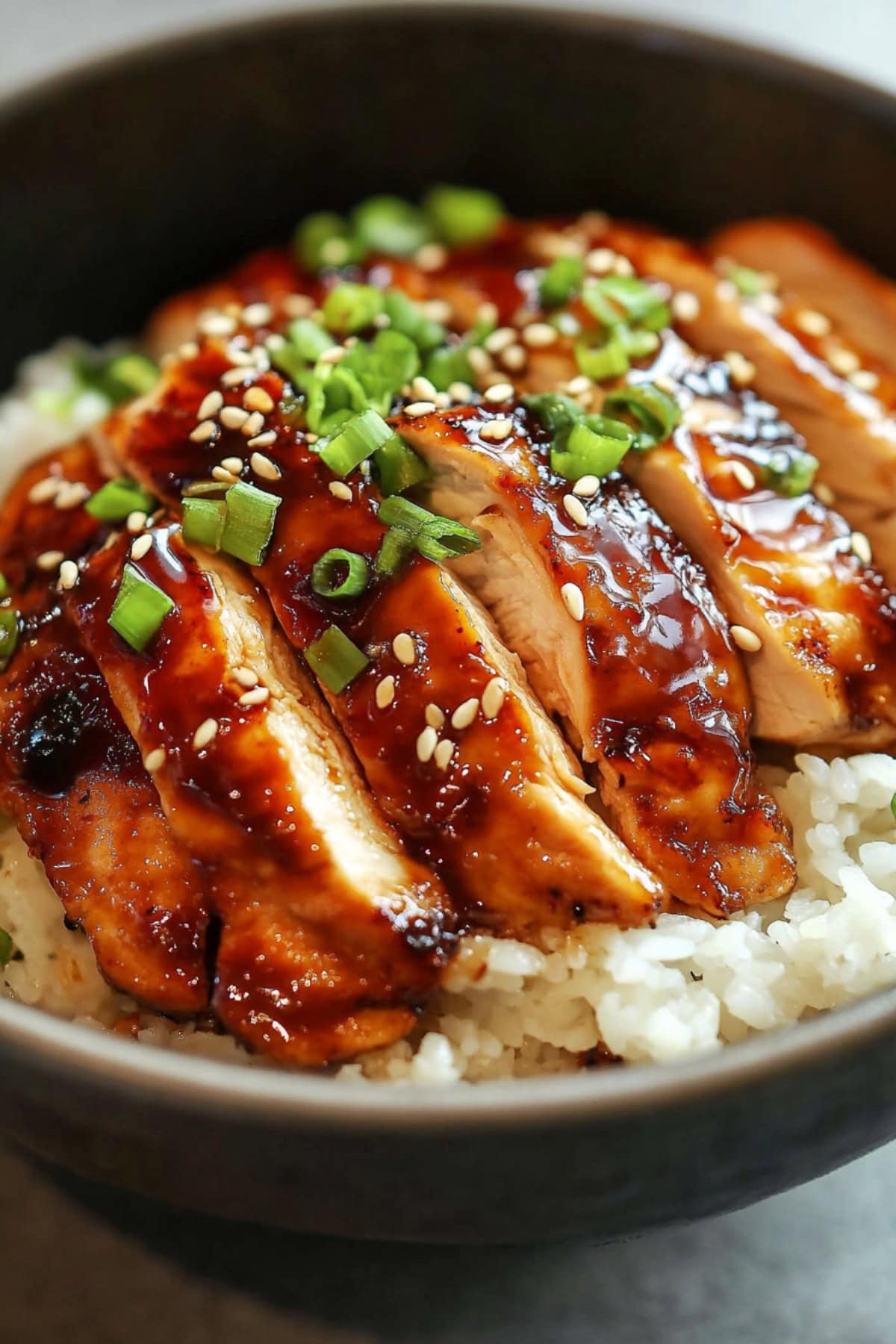 Sliced teriyaki chicken served over white rice in a black bowl.