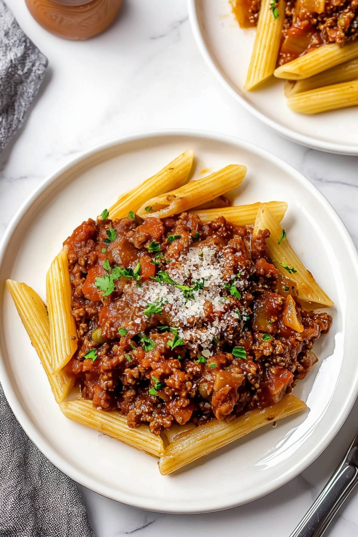 Penne bolognese with ground beef, grated Parmesan and fresh herbs on a white plate, top view