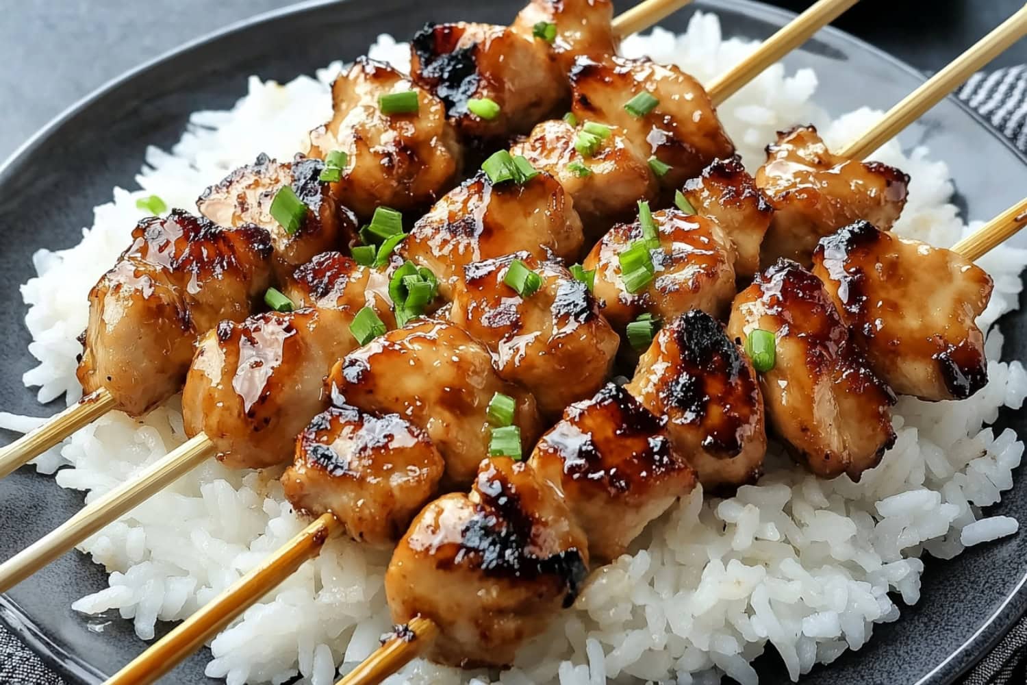 Golden-brown teriyaki chicken skewers, garnished with chopped green onions, and served with rice in a plate.