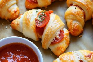 Crescent rolls with pepperoni filling inside sitting on a parchment paper with pizza sauce dip in a small bowl.